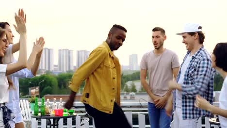 Joyful-African-American-man-in-trendy-clothes-is-dancing-with-friends-multiracial-group-having-fun-at-party-on-roof-on-summer-day.-Leisure,-joy-and-friendship-concept.