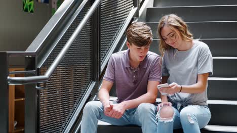 Männliche-und-weibliche-Schülerinnen-und-Schüler-sitzen-auf-Treppen,-die-Nachricht-auf-dem-Handy-betrachten