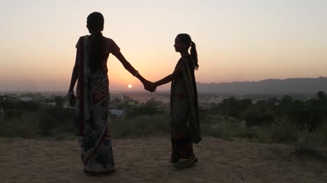 Two-women-stand-hold-hands-at-sunset-beautiful-vantage-point-romantic-setting-dawn-high-wide-panoramic-surreal-lovers-in-traditional-dress-silhouette-in-Rajasthan-India-handheld-from-behind-medium-two