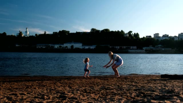 Beautiful-blonde-mom-and-daughter-run-towards-each-other-on-the-river-Bank-at-sunset.