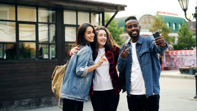 Glücklicher-afroamerikanischer-Kerl-nimmt-Selfie-mit-Freunden-schöne-Kaukasische-Mädchen-stehen-in-Straße-posieren-und-Smartphone-während-der-Reise-im-Ausland-zu-halten.