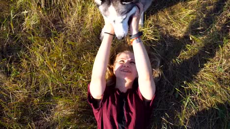 Top-view-of-young-girl-with-blonde-hair-lying-on-grass-at-field-and-stroking-her-husky-dog.-Beautiful-woman-spending-time-together-with-her-pet-at-nature.-Love-and-friendship-with-domestic-animal.