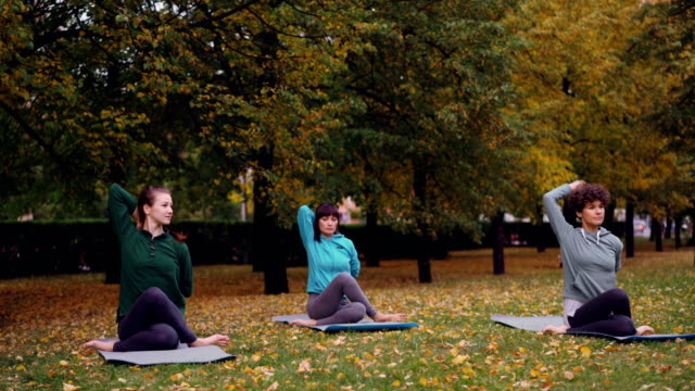 Relaxed-females-are-sitting-on-yoga-mats-in-Cow-Face-pose-and-stretching-arms-while-professional-instructor-is-speaking-explaining-correct-position.