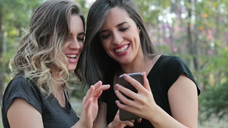 Two-girlfriends-holding-cellphone-laughing-and-smiling-together.-Latin-women-holding-smartphone-laugh-at-content-they-are-watching