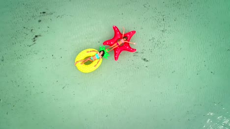 Luftaufnahme-von-zwei-Mädchen-auf-Luftmatratze-im-transparenten-Meer-schwimmende-halten-einander.
