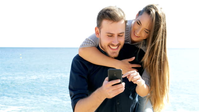 Happy-couple-joking-and-using-phone-on-the-beach