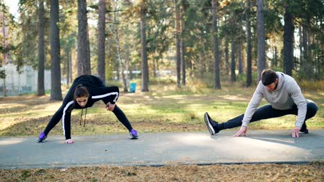 Young-handsome-man-and-female-friend-are-training-in-park-together-stretching-legs-on-warm-autumn-day-wearing-trendy-tracksuits.-People,-sports-and-leisure-concept.