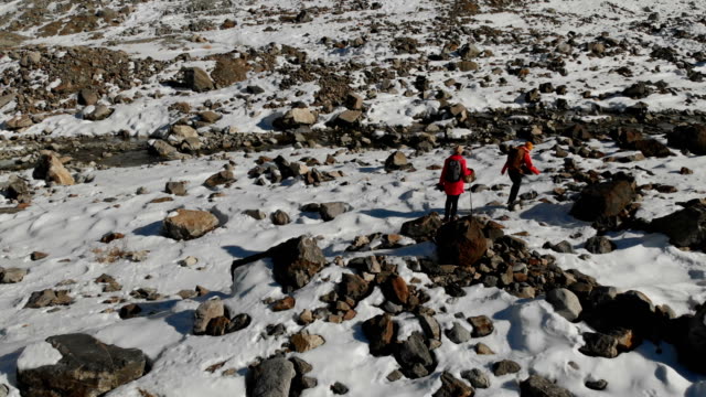Luftaufnahme-von-zwei-Mädchen-Reisenden-mit-Rucksäcken-und-Kameras-gehen-durch-den-Schnee-und-Steinen-auf-den-Gletscher-zwischen-den-epischen-Felsen-in-den-Bergen.-Mädchen-Fotografen-mit-ihren-Kameras-bei-Sonnenuntergang