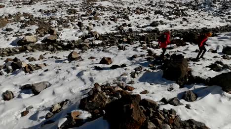 Luftaufnahme-von-zwei-Mädchen-Reisenden-mit-Rucksäcken-und-Kameras-gehen-durch-den-Schnee-und-Steinen-auf-den-Gletscher-zwischen-den-epischen-Felsen-in-den-Bergen.-Mädchen-Fotografen-mit-ihren-Kameras-bei-Sonnenuntergang