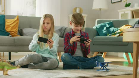 Zu-Hause-auf-einem-Teppich-sitzen:-niedliche-kleine-Mädchen-und-süße-junge-Videospiel-in-wettbewerbsfähige-auf-zwei-Smartphones,-hielt-sie-im-horizontalen-Modus-\"Querformat\".-In-Zeitlupe.