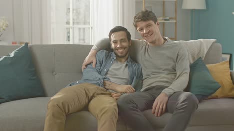 Pareja-Gay-masculino-atractivo-lindo-sentarse-juntos-en-un-sofá-en-casa.-Novios-son-Hugging-y-abrazándose-unos-a-otros.-Ellos-son-feliz-y-sonriente.-Son-vestidos-casualmente-y-sala-tiene-un-Interior-moderno.
