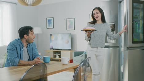 Beautiful-Young-Couple-Talking-in-a-Kitchen-at-Home.-Man-is-Sitting-at-a-Table,-Girl-Takes-a-Cake-out-of-the-Fridge.-Thay-are-Happy-and-Laugh.-Room-has-Modern-Interior.