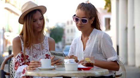 Two-female-friends-read-a-guidebook-outside-a-cafe,-Ibiza