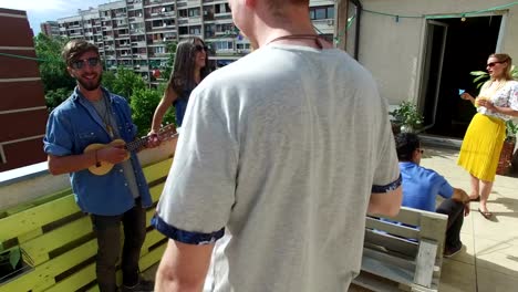 Mann-spielt-Ukulele,-während-er-mit-Freunden-auf-dem-Dach-hängt
