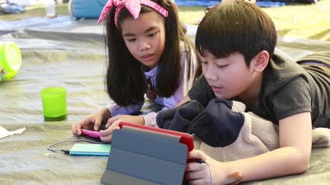 Feliz-chico-asiático-y-niña-jugando-teléfono-inteligente-y-tableta-en-la-zona-de-acampada