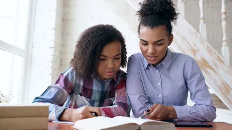 Teenage-lockig-gemischt-Rasse-junge-Mädchen-sitzen-am-Tisch-konzentriert-konzentriert-Lektionen-und-ihre-ältere-Schwester-hilft-ihr-zu-studieren