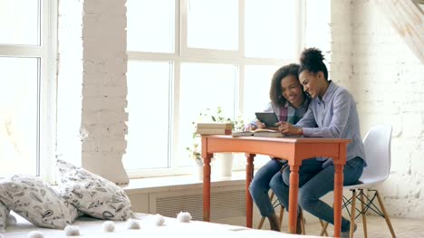 Zwei-attraktive-lockige-gemischte-Rasse-junge-Mädchen-sitzen-am-Tisch-haben-Spaß-beim-Lernen-Unterricht-und-mit-Tablet-zu-Hause