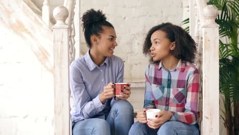 Zwei-afrikanische-amerikanische-lockige-Mädchen-Sistres-sitzen-auf-Treppen-haben-Spaß-lachen-und-chatten-zusammen-zu-Hause