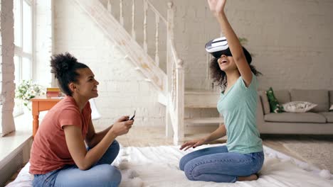 Mixed-raced-woman-using-virtual-reality-glasses-while-her-friend-holding-digital-tablet-computer.-Girlfriends-play-video-game-3d-technology-concept-at-home