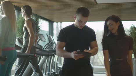 Hombre-y-mujer-atléticas-están-usando-una-tableta-junto-a-las-cintas-de-correr-en-un-gimnasio-deportivo.