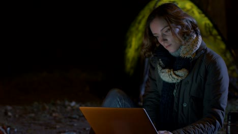 Brunette-Mädchen-benutzt-nachts-einen-Laptop-neben-einem-Lagerfeuer-mit-einem-Zelt-im-Hintergrund.