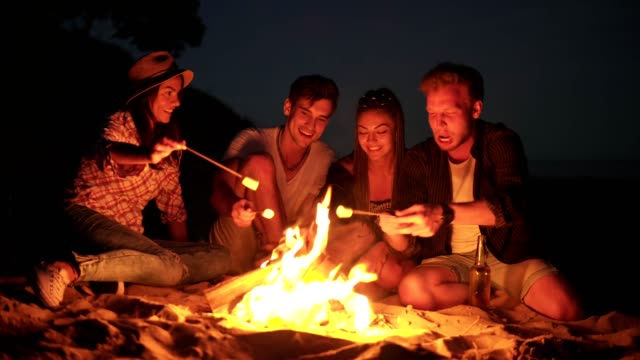 Jóvenes-amigos-alegres-sentado-junto-al-fuego-en-la-playa-por-la-noche,-cocinar-melcocha-en-palillos-juntos.-Rodada-en-4k