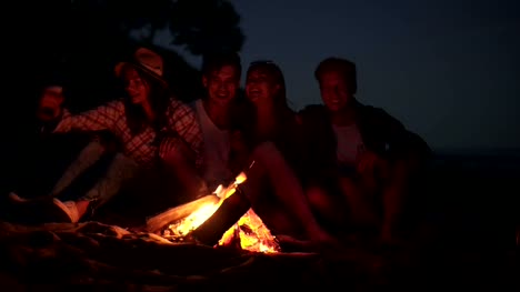 Young-cheerful-friends-sitting-by-the-fire-on-the-beach-in-the-evening,-singing-songs-and-drinking-beer-together.-Shot-in-4k