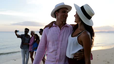 Paar-zu-umarmen,-zu-küssen-am-Strand-mit-Blick-auf-Sonnenuntergang-über-Volksgruppe-sprechen-Selfie-Foto,-Touristen-Mann-und-Frau-im-Sommer-Urlaub