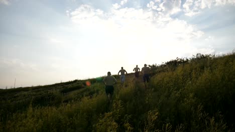 Gruppe-junger-Männer,-die-den-grünen-Hügel-hinunter-über-blauer-Himmel-mit-Sonne-Streulicht-im-Hintergrund-laufen.-Männliche-Athleten-ist-Joggen-in-der-Natur-bei-Sonnenuntergang.-Kufen-bergab-Sport-im-Freien-bei-Sonnenaufgang.-Slow-motion