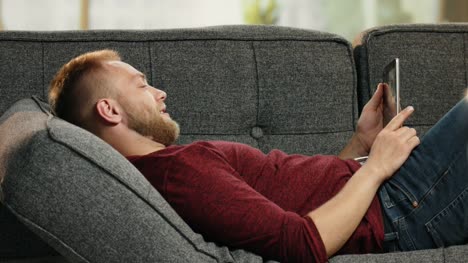 Atractivo-joven-acostado-en-el-sofá-leyendo-noticias-divertidas-sobre-tableta