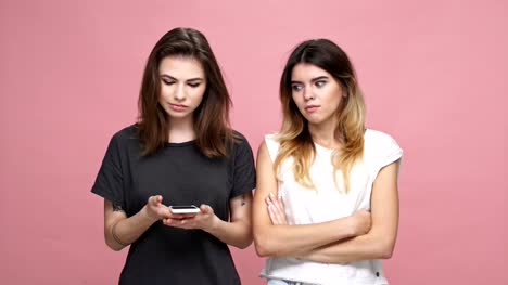 Two-young-casual-girls-looking-at-mobile-phone-and-getting-upset-isolated-over-pink-background