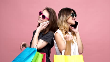 Two-smiling-pretty-women-in-sunglasses-standing-back-to-back-with-shopping-bags-and-talking-on-mobile-phone-isolated-over-pink-background
