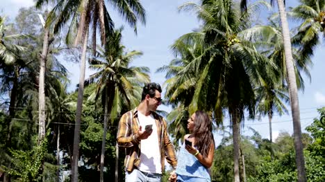 Couple-Walk-Using-Cell-Smart-Phone-Under-Palm-Trees,-Man-Calling-Talk,-Smiling-Man-And-Woman-Online-Communication