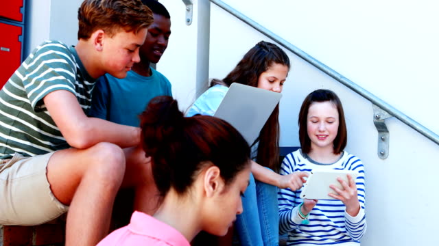 Group-of-smiling-school-friends-on-staircase-using-laptop-and-digital-tablet