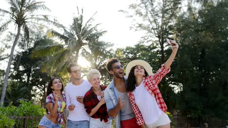 Grupo-de-personas-tomar-foto-Selfie-en-celulares-inteligentes-teléfonos-hablando-felices-hombres-y-mujeres-sobre-palmeras-tropicales