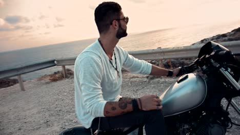 Hombre-joven-rebelde-sentado-en-la-motocicleta-y-mirando-la-puesta-del-sol