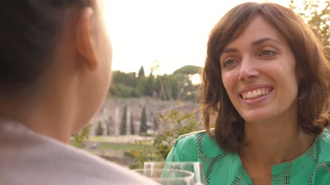 Zwei-glückliche-junge-Frau-Touristen-sitzen-am-Tisch-der-eine-Bar-Restaurant-vor-dem-Kolosseum-in-Rom-trinken-und-mit-einem-Glas-italienischen-Rotwein.-Stilvolle-buntes-Kleid-an-einem-Sommertag-bei-Sonnenuntergang
