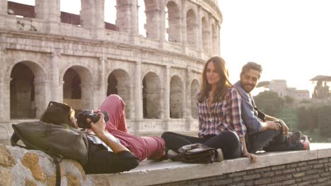 Three-young-friends-tourists-sitting-lying-in-front-of-colosseum-in-rome-at-sunset-taking-pictures-photos-with-dslr-camera-backpacks-sunglasses-happy-beautiful-girl-long-hair-slow-motion