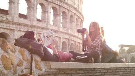 Mochilas-de-tres-turistas-jóvenes-amigos-sentados-mentira-delante-de-Coliseo-de-Roma-al-atardecer-tomando-fotos-fotos-con-dslr-cámara-gafas-de-sol-feliz-hermosa-chica-de-cabello-largo-lenta