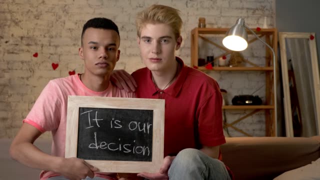 A-sad-international-gay-couple-is-sitting-on-the-couch-and-holding-a-sign.-It-is-our-decision.-Look-at-the-camera.-Home-comfort-on-the-background.-60-fps