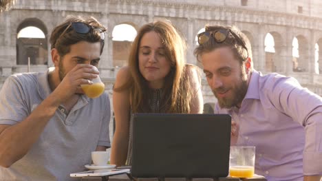 Three-happy-friends-having-video-call-with-laptop-sitting-at-bar-restaurant-table-in-front-of-colosseum-in-rome-at-sunset-waving-hands-giving-thumbs-up