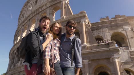 Three-young-friends-tourists-standing-on-pedestal-in-front-of-colosseum-in-rome-taking-funny-hilarious-pictures-posing-with-backpacks-sunglasses-happy-beautiful-girl-long-hair-slow-motion