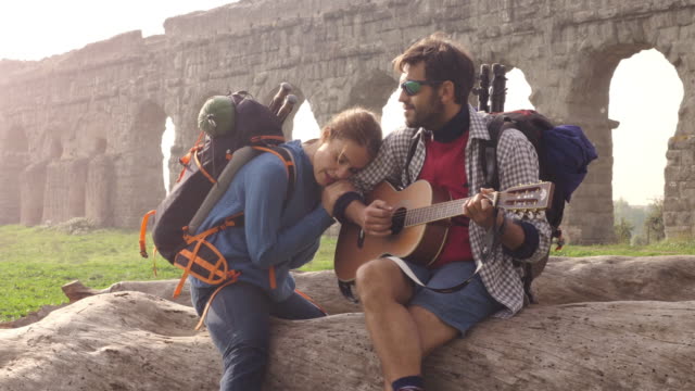 Glückliches-junges-Paar-Backpacker-Touristen-sitzen-auf-einem-Log-Stamm-spielen-Gitarre-singen-vor-Ruinen-der-antiken-römischen-Aquädukt-in-romantischen-Parco-Degli-Acquedotti-Park-in-Rom-bei-Sonnenaufgang-Schlafsack-Slow-motion