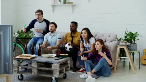 Group-of-young-friends-watching-football-game-on-TV-together-eating-snacks-and-drinking-beer-at-home.-Men-are-happy-with-their-team-winning-but-girls-disappointed
