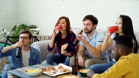 Happy-friends-have-party-talking-and-eating-pizza.-They-clink-bottles-sitting-on-sofa-at-home