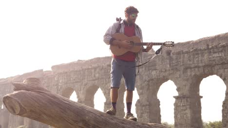 Viajero-de-aventurero-joven-de-pie-sobre-un-tronco-de-registro-jugando-canto-guitarra-frente-antiguo-acueducto-romano-ruinas-en-parco-degli-acquedotti-Parque-de-Roma-en-la-lenta-salida-del-sol