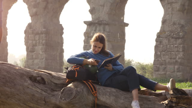Happy-young-couple-backpackers-tourists-on-a-log-trunk-playing-guitar-singing-reading-book-in-front-of-ancient-roman-aqueduct-ruins-in-romantic-parco-degli-acquedotti-park-in-rome-at-sunrise-slow-motion