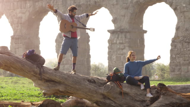 Happy-young-couple-backpackers-tourists-on-a-log-trunk-taking-selfies-photos-with-smartphone-in-front-of-ancient-roman-aqueduct-ruins-in-romantic-parco-degli-acquedotti-park-in-rome-at-misty-sunrise-slow-motion-TRIPOD