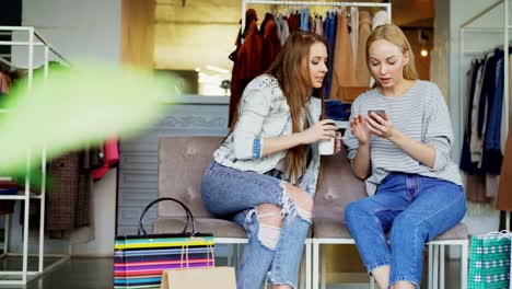Female-friends-talking-and-checking-smartphone-while-sitting-is-shopping-center.-Women's-clothing-and-paperbags-in-background.