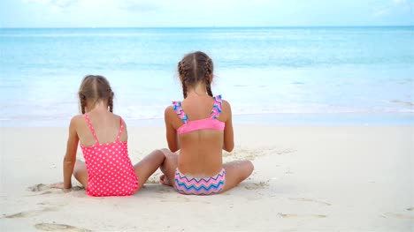 Adorable-little-girls-playing-with-sand-on-the-beach.-Kids-sitting-in-shallow-water-and-making-a-sandcastle